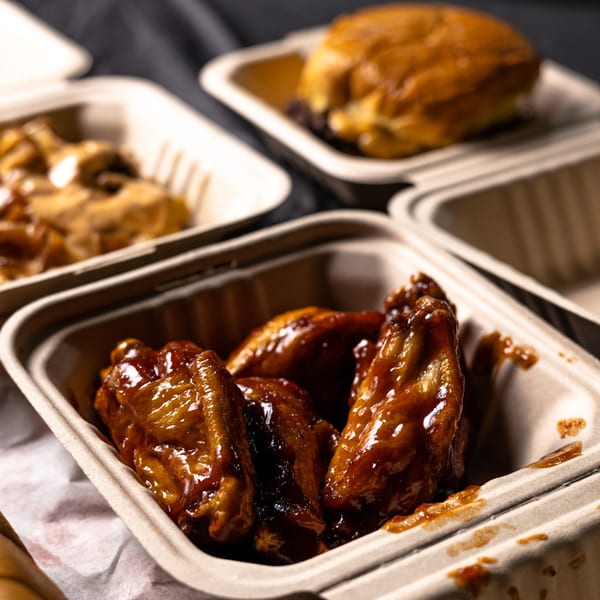 Close-up of Bricked Burgers' BBQ chicken wings in a takeout container, with a burger in the background.
