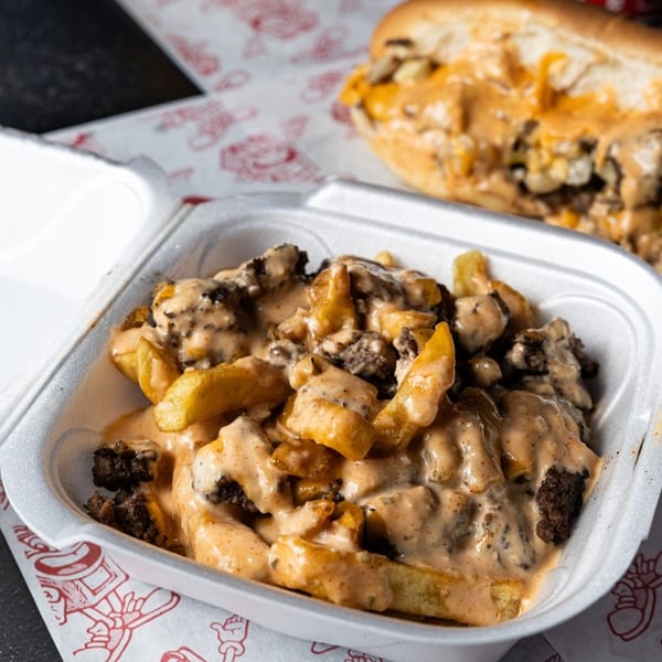 Bricked Burgers' loaded fries with cheese sauce and ground beef, with a cheesesteak in the background.